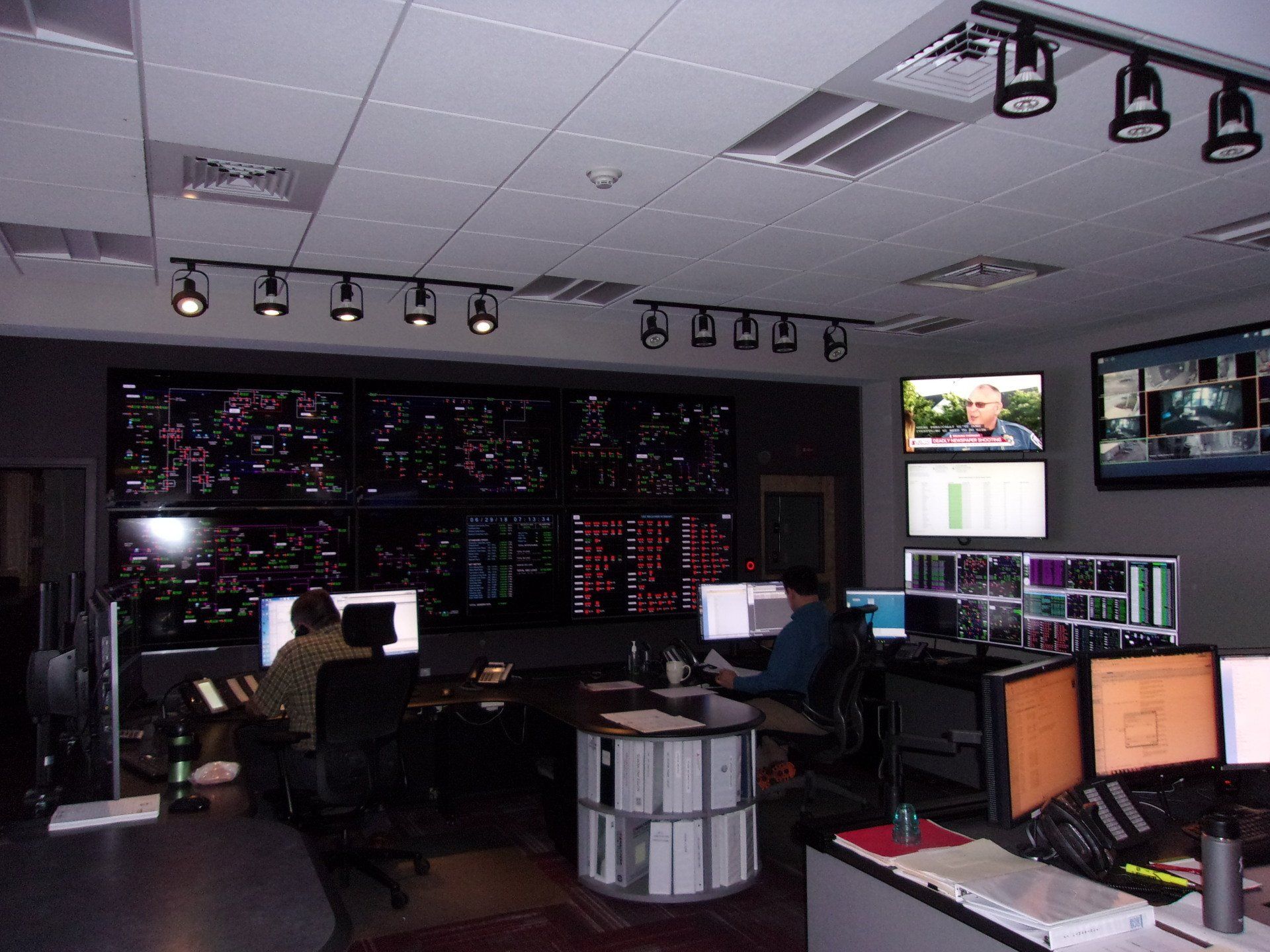 Control room with multiple monitors, two operators at consoles, and wall displays showing data and video feeds.