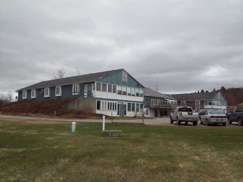 Large blue building with white trim on a grassy lot under a cloudy sky, with parked cars in front