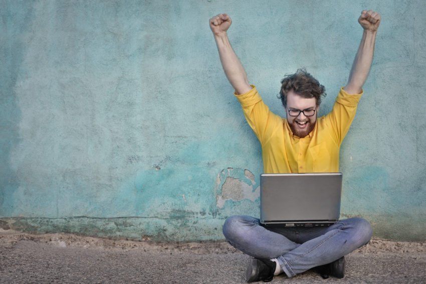 Een man met opgeheven armen, glimlachend naar zijn laptop, zit met gekruiste benen tegen een turquoise muur.