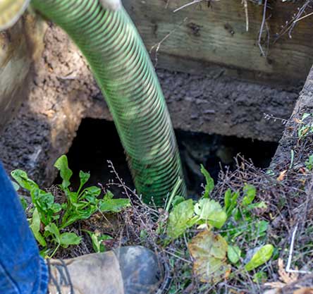 Green hose entering an open septic tank. Mud, grass, and part of a boot visible.