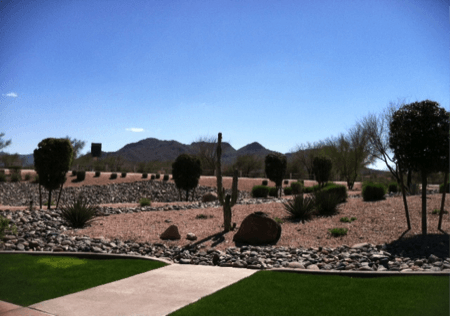 Desert landscape with stone-filled beds, green grass, and mountains under a bright blue sky.