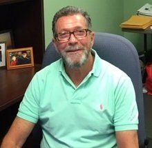 Man in a light green polo shirt and glasses smiles at the camera in an office setting.