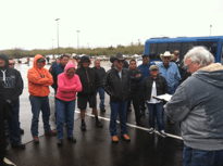 Group of people listening to a man, standing in a parking lot in front of a blue bus, on a rainy day.