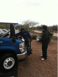 Two people by a blue van; one working under the hood, the other holding a clipboard.