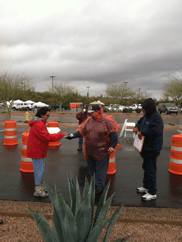 People in rain gear handing out items near orange cones and a parking lot. Overcast sky.