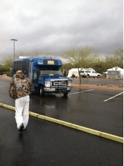 Man in camouflage walks toward a blue bus in a wet parking lot; cloudy day.