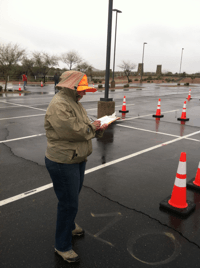 Person in an orange hat and jacket standing outside, writing on a clipboard, near traffic cones on a wet parking lot.