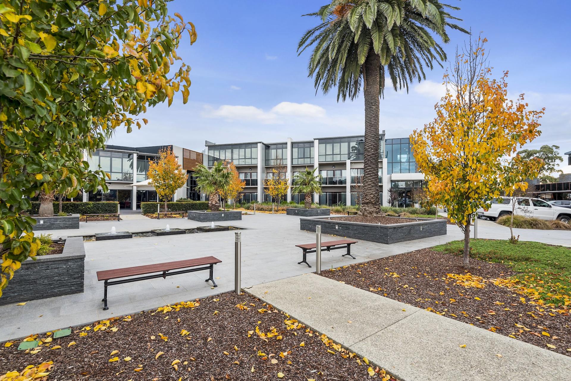 A modern apartment building with benches, trees, and a central walkway. Autumn leaves and blue sky.