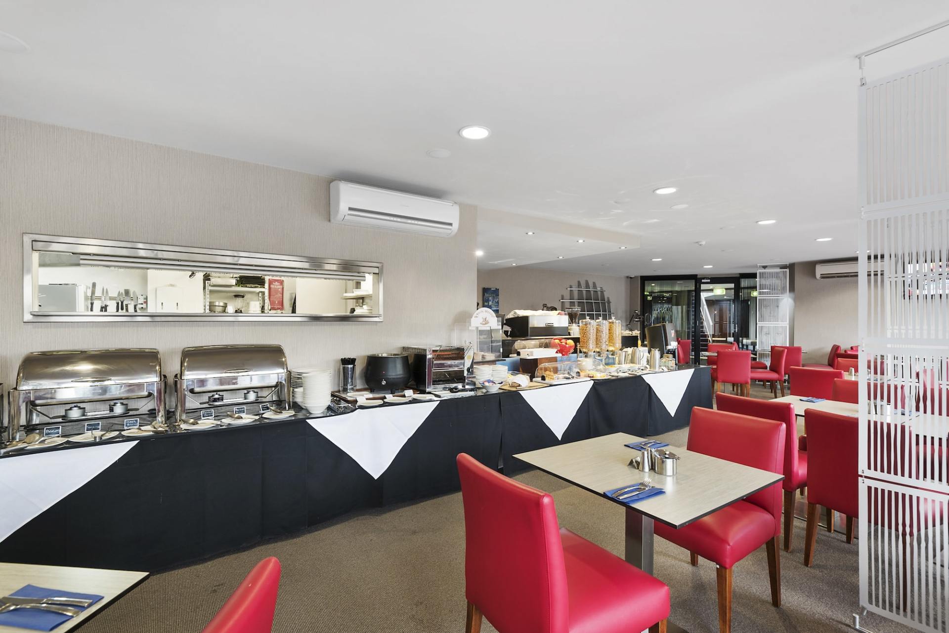 Breakfast buffet in a hotel dining area with red chairs and white tables. Food items displayed on a buffet counter.