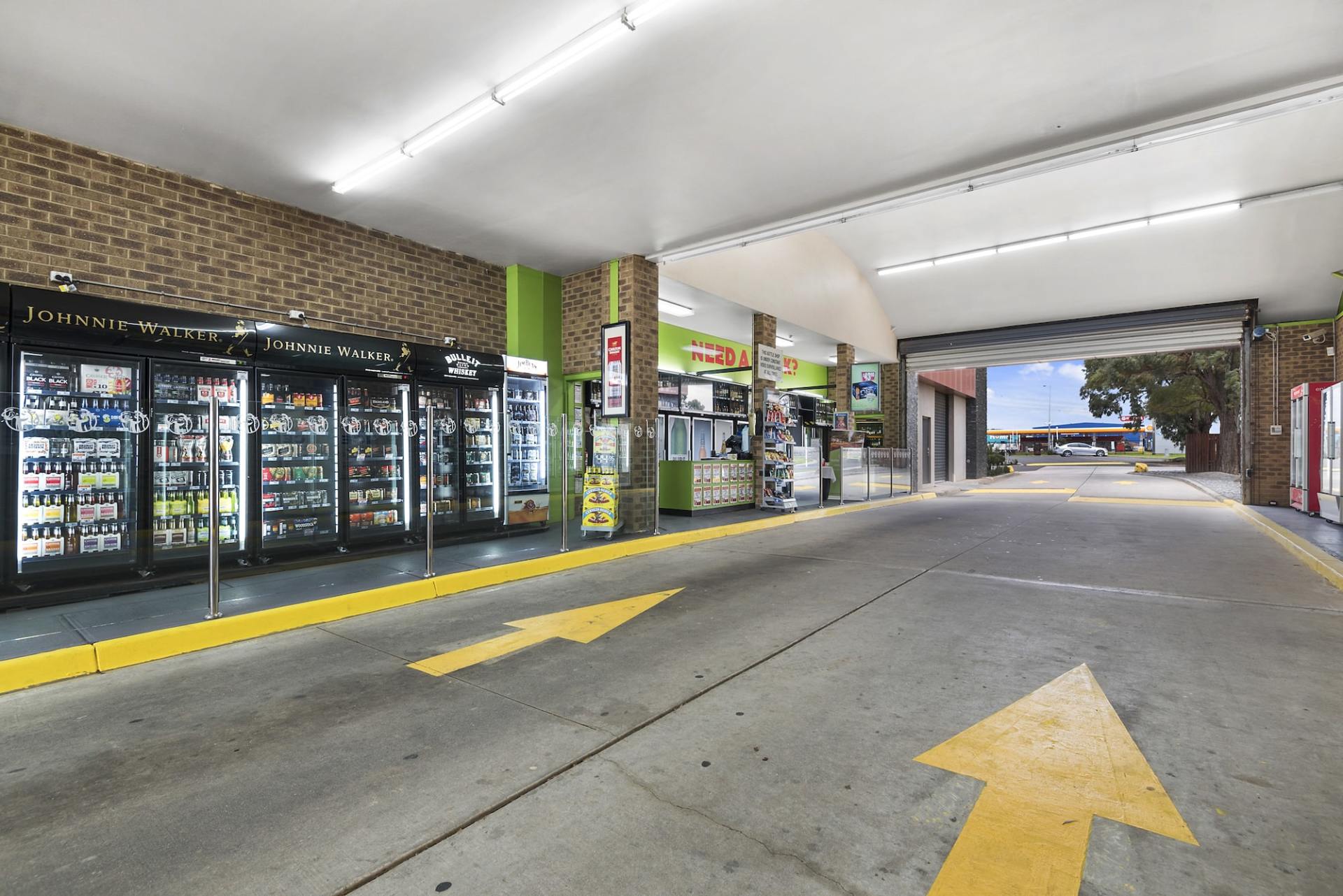 A convenience store entrance with yellow arrows on concrete, refrigerators, and a small shop.