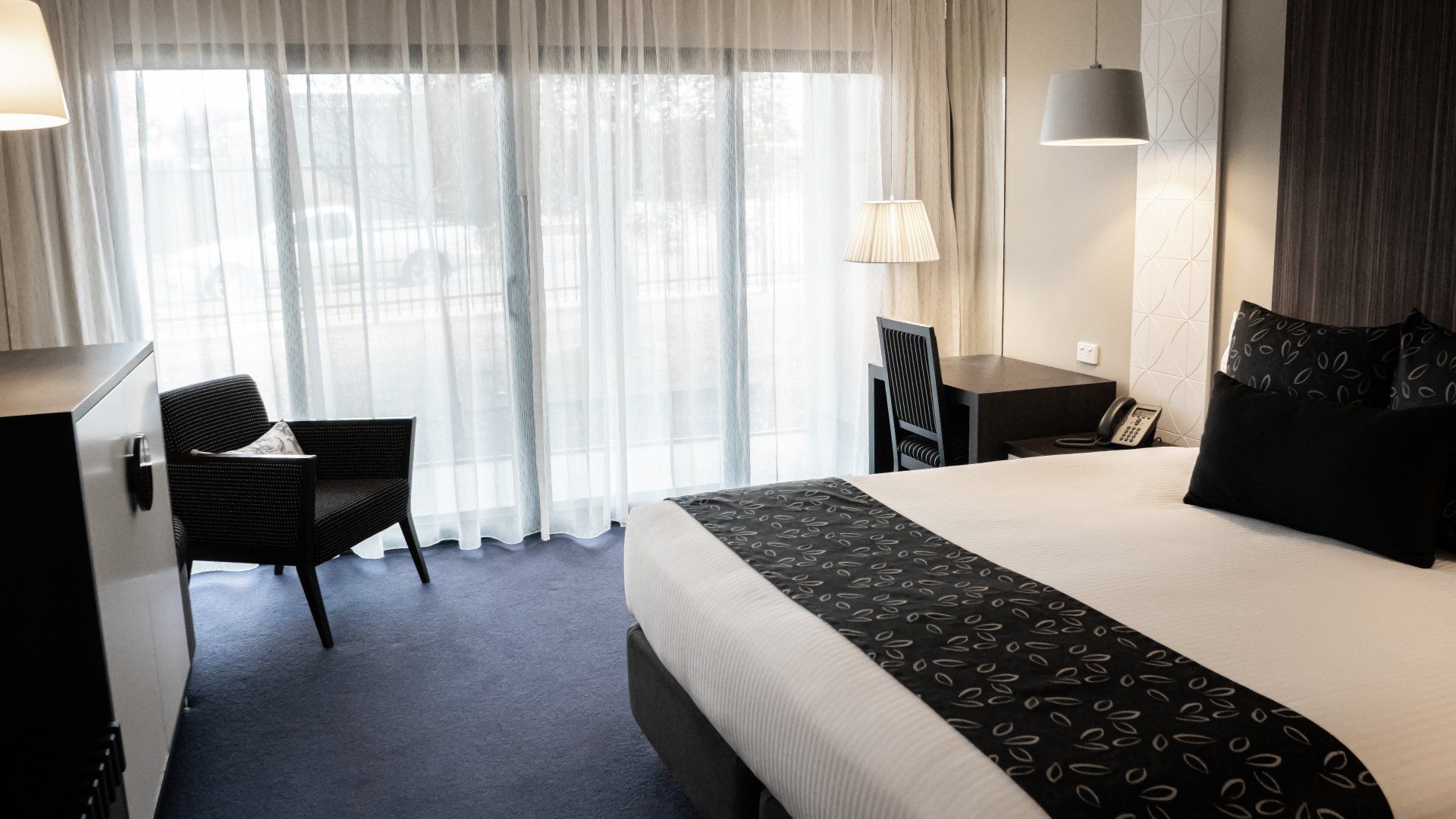 Hotel room with bed, desk, chair, and sheer curtains.