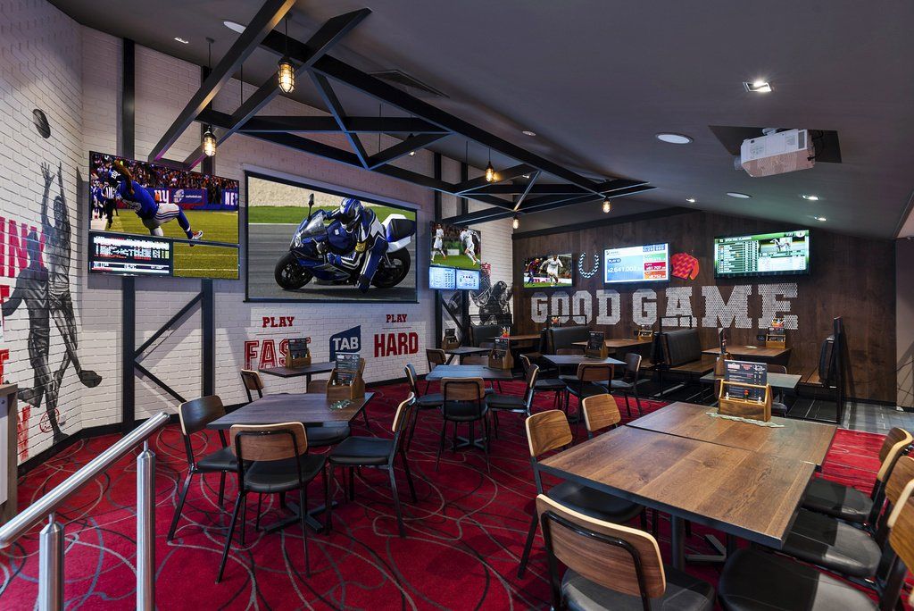 Bar interior with tables, chairs, and TVs showing sports; red carpet, dark wood accents.