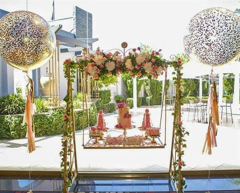Pink floral swing decorated for a party with a cake, surrounded by balloons and greenery.