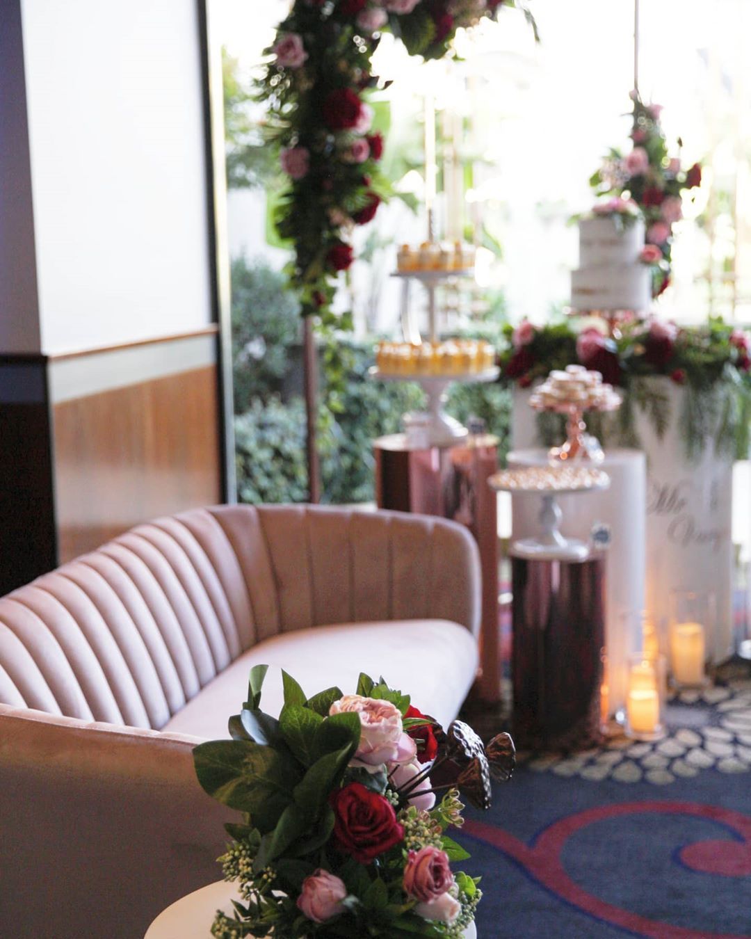 Romantic wedding reception: pink couch, floral arch, cake, cupcakes on rose gold pedestals.