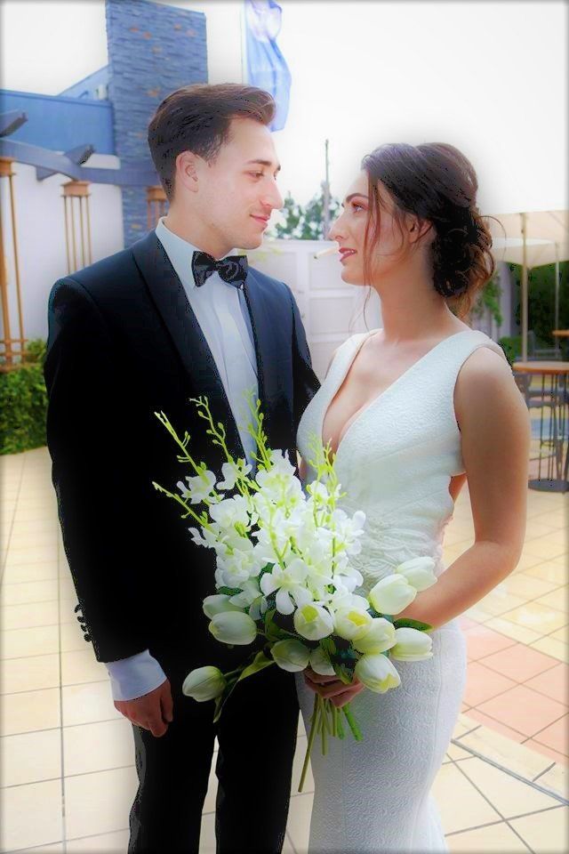 Newlyweds gaze at each other. Bride in white dress holds bouquet, groom in black tux. Outdoor setting.