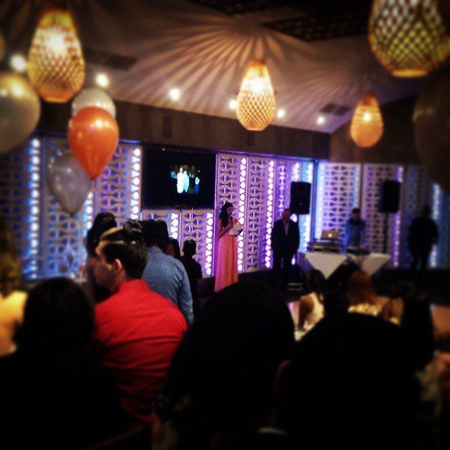 People at an event with a speaker on stage. Orange and gold lanterns hang from the ceiling.