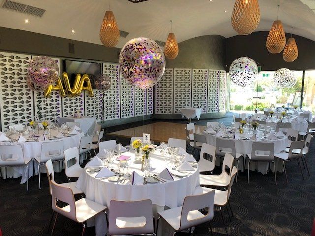 Formal event room with round tables, white chairs, balloons, and hanging lights.
