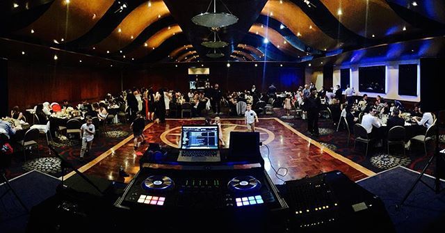 DJ setup at a formal event; people dance on a wooden floor, surrounded by tables in a large ballroom.