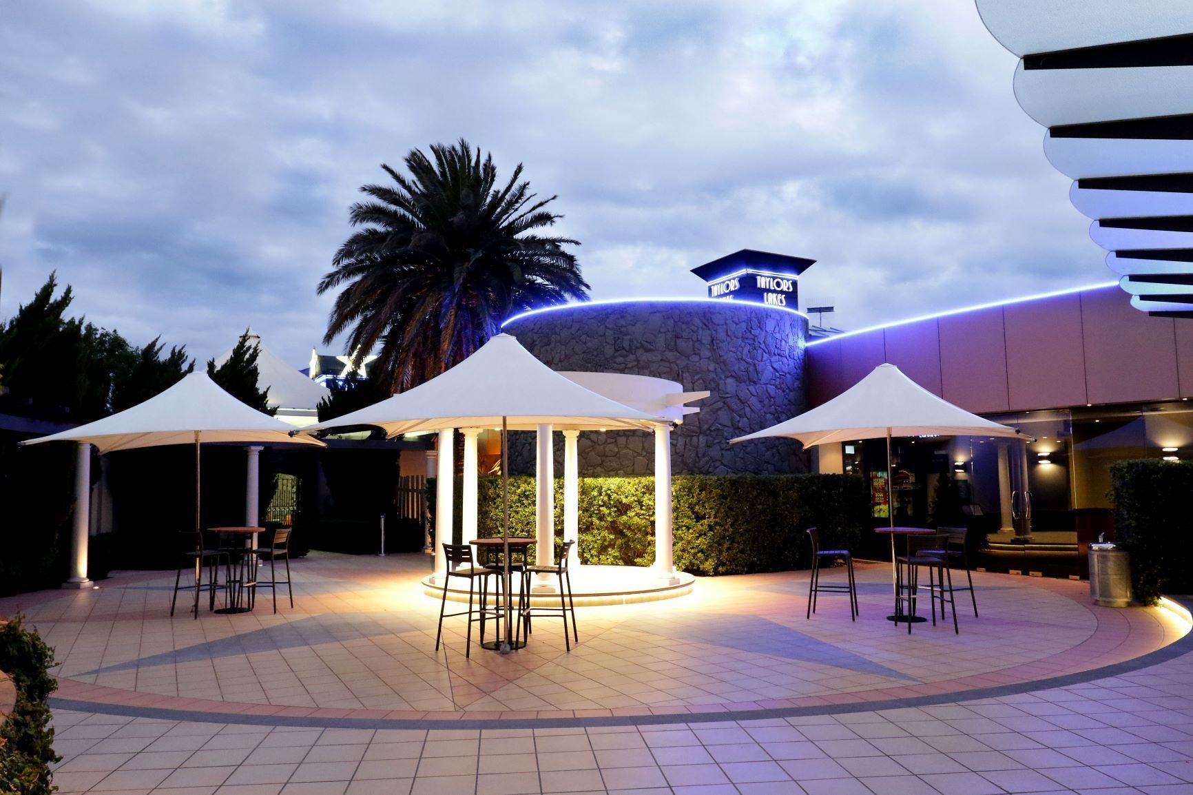 Outdoor patio with three white umbrellas over tables, evening lighting.