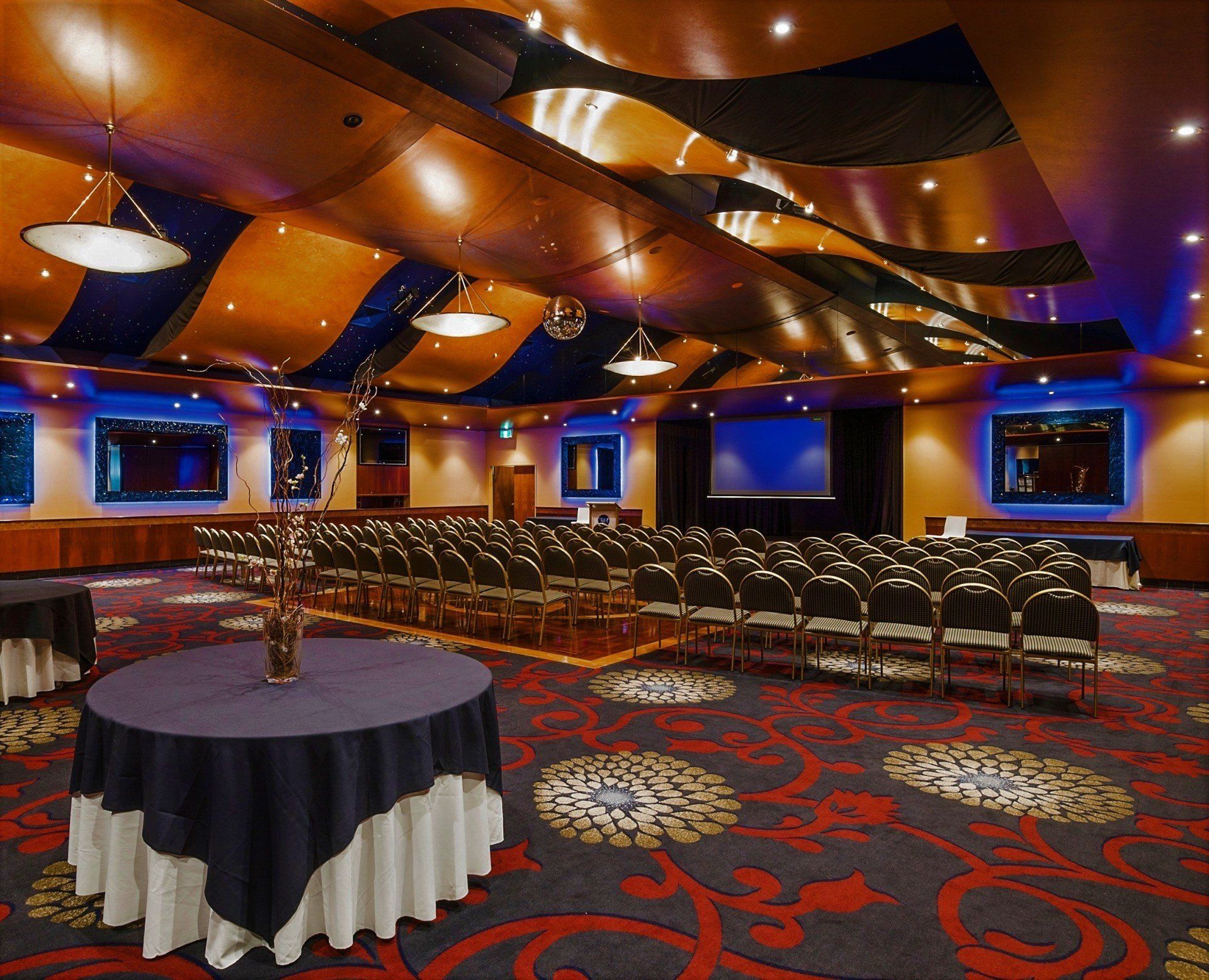 Auditorium with rows of chairs, stage, round tables, patterned carpet, and decorative ceiling.