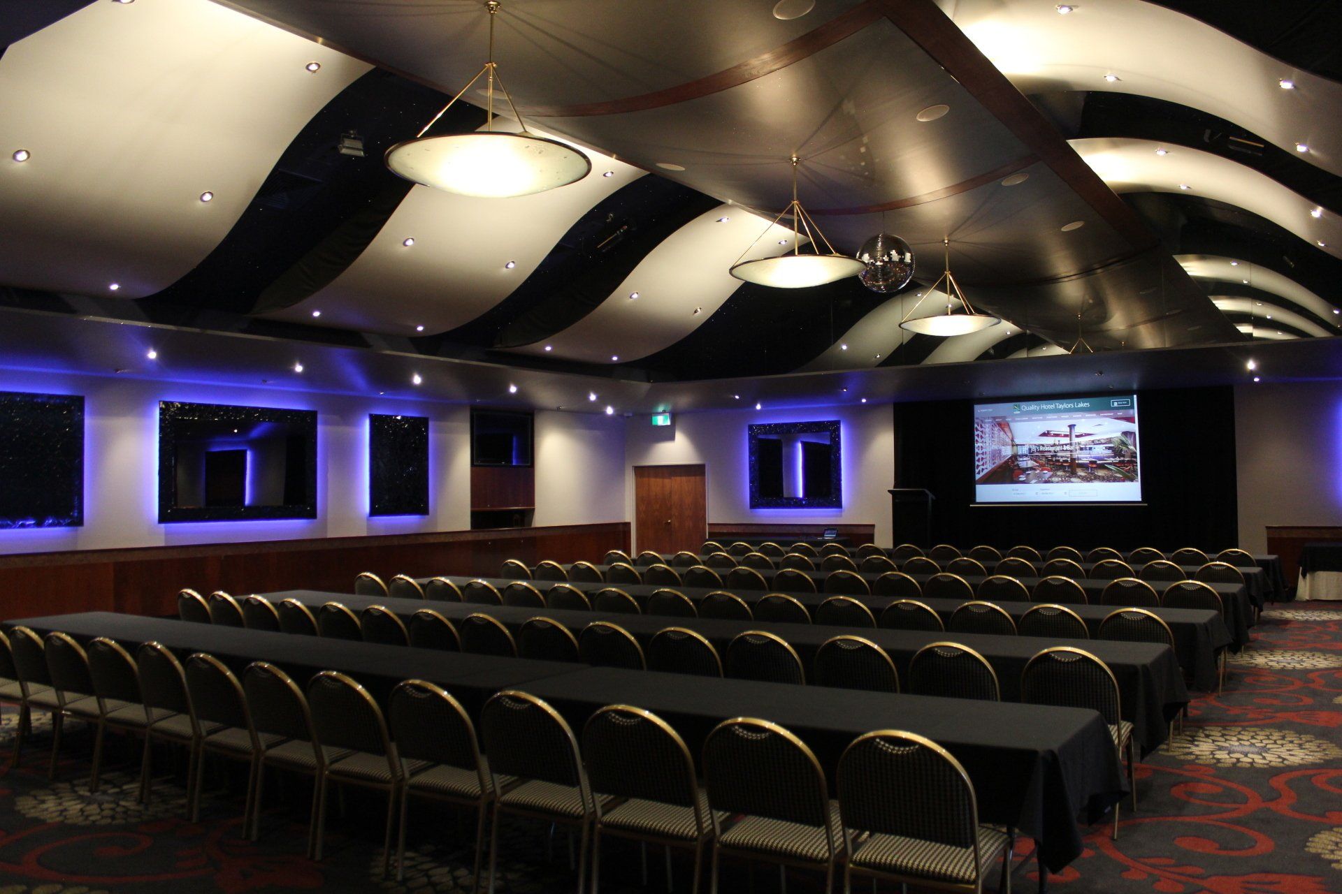 Auditorium with rows of tables and chairs, screen, and decorative black and white ceiling.