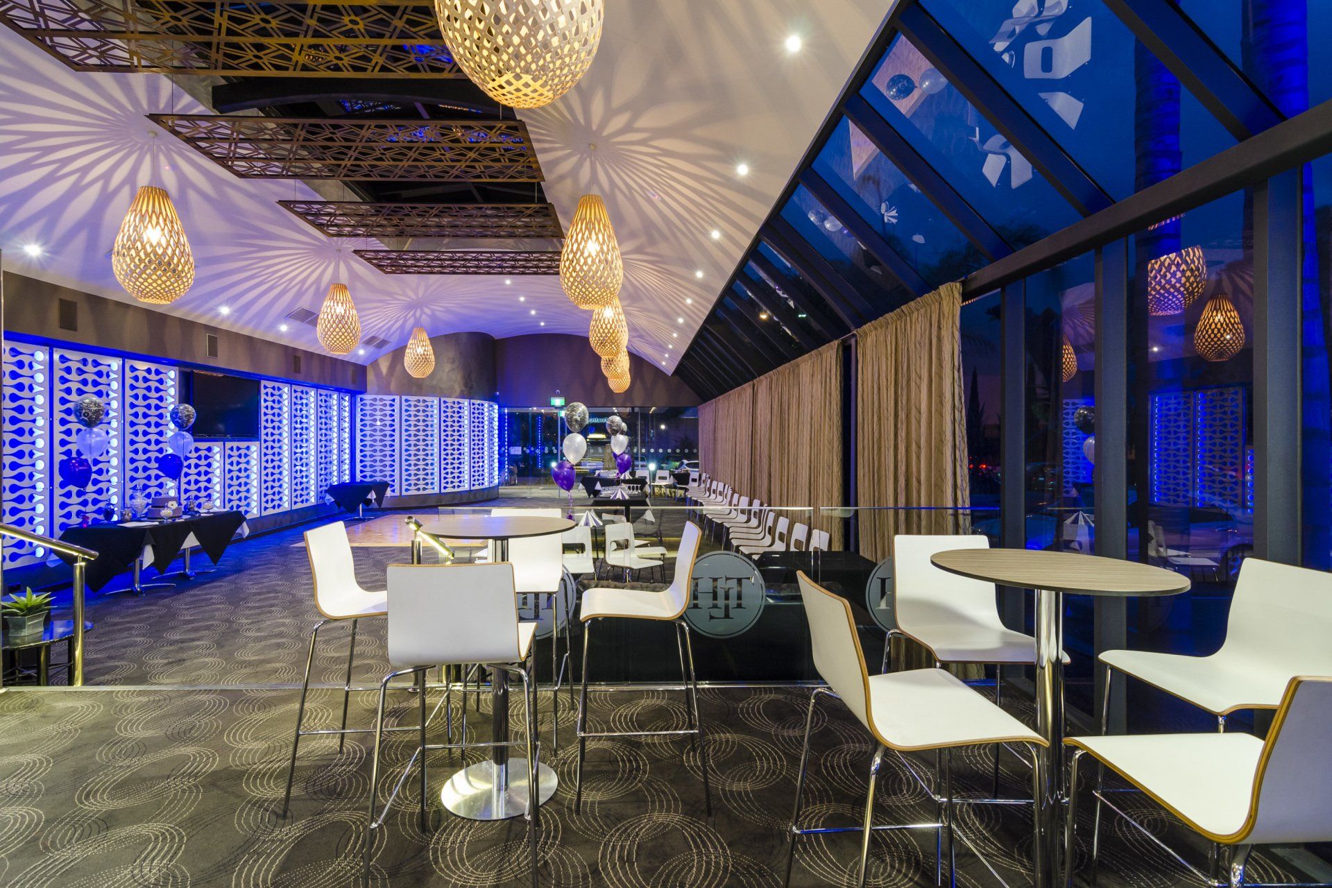 Modern bar interior with white chairs, round tables, ornate ceiling lights, and blue-lit wall.
