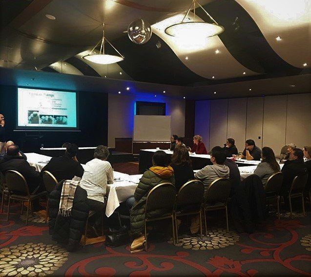 People seated around tables in a room, attending a presentation projected on a screen.