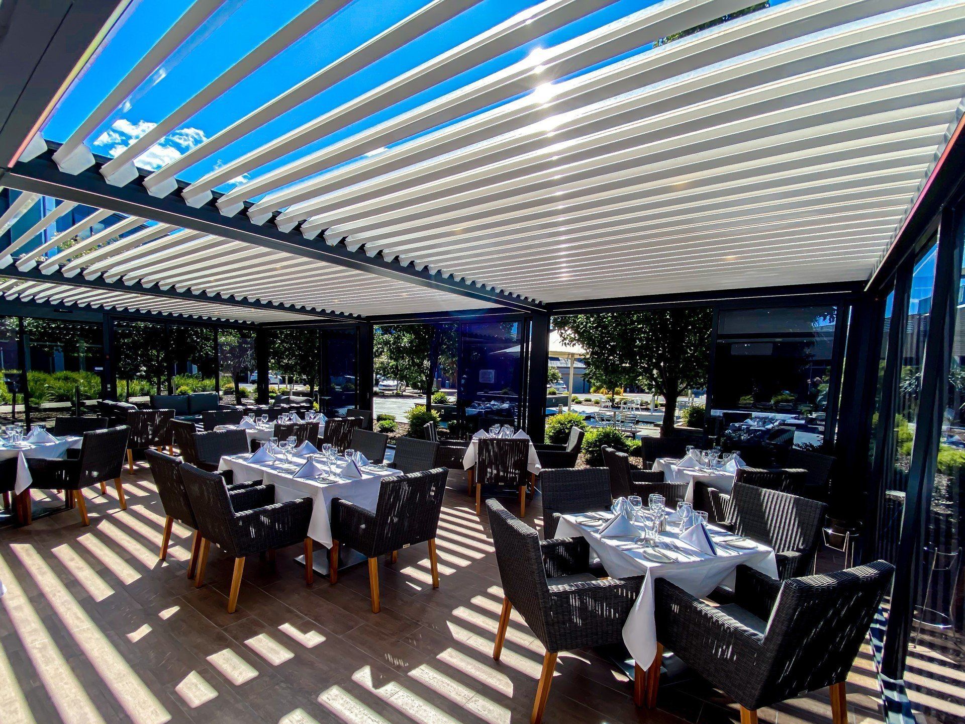 Outdoor restaurant with tables set for dining, under a slatted, adjustable roof. Sunlight casts striped shadows.