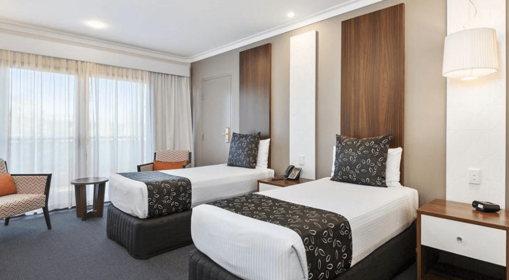 Two twin beds in a modern hotel room with dark wood headboards, a window, and a small sitting area.