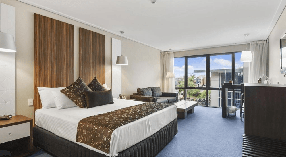 Hotel room with a king bed, sofa, and large window overlooking city.