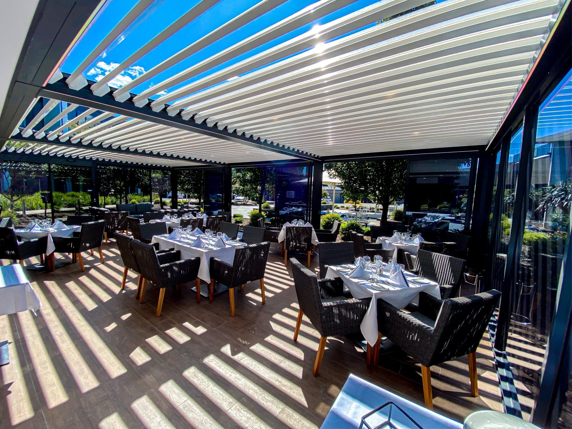 Outdoor restaurant dining area with white tablecloths, black chairs, and a slatted roof, on a sunny day.