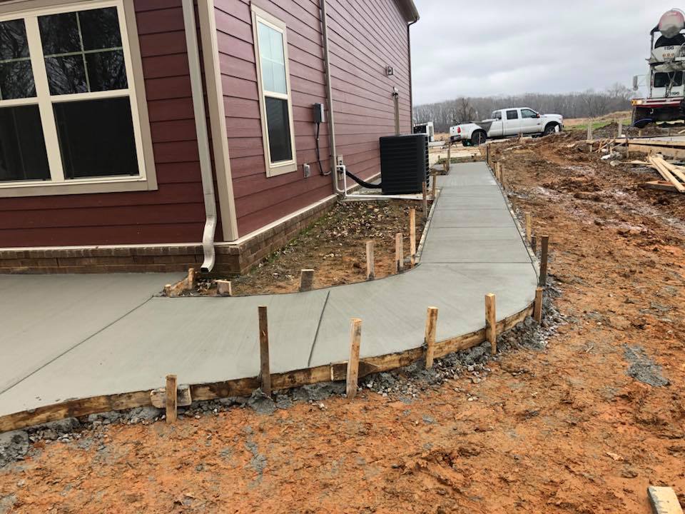 Concrete walkway beside a red house under construction, with exposed dirt and wooden stakes along the edge