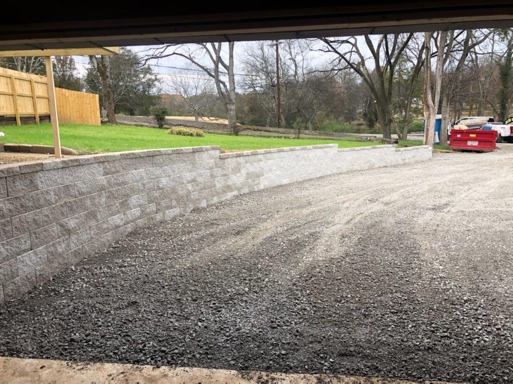 Gravel driveway under an overpass, with trees, grass, and a red car in the distance.