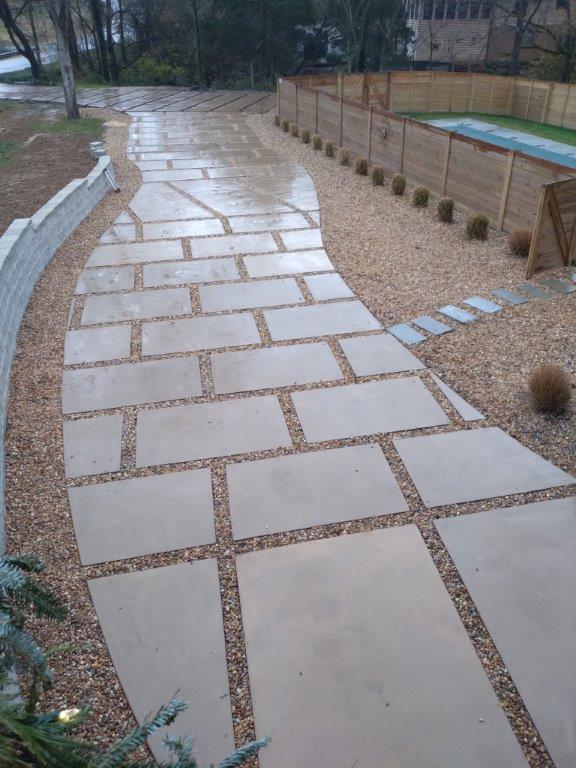 Wet stone walkway beside a fenced pool in a garden, with pebble borders and light gray paving slabs