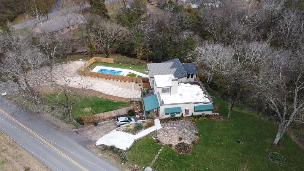 Aerial view of a white house with a backyard pool beside a road and wooded yard.