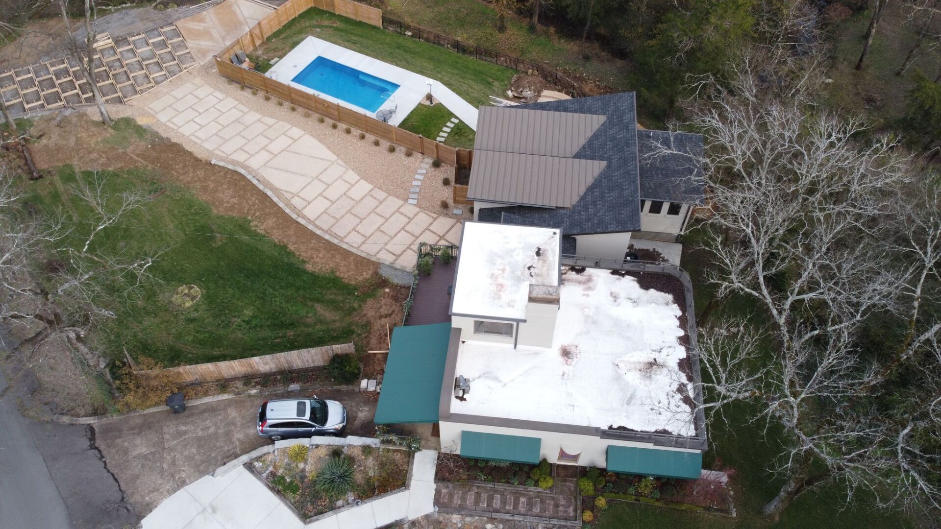 Aerial view of a house with a white roof, swimming pool, patio, and parked car beside trees.