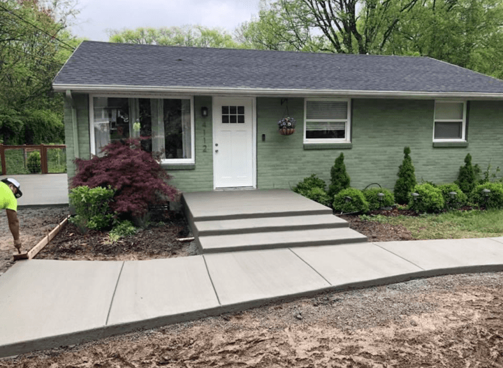 Small green house with white trim, front steps, walkway, and landscaped shrubs.