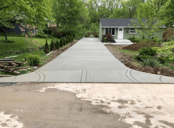 Wide concrete driveway leading to a small house surrounded by lush green trees and shrubs.
