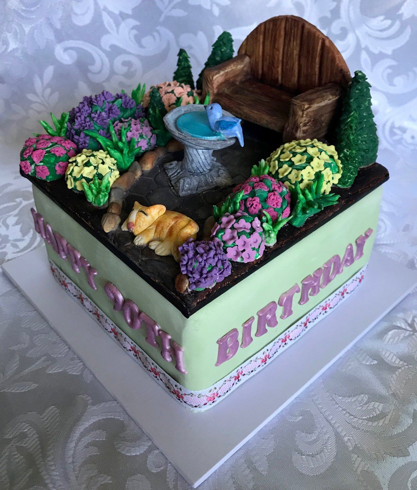 Square Garden themed birthday cake with garden chair, bird bath, flowering bushes and little cat.