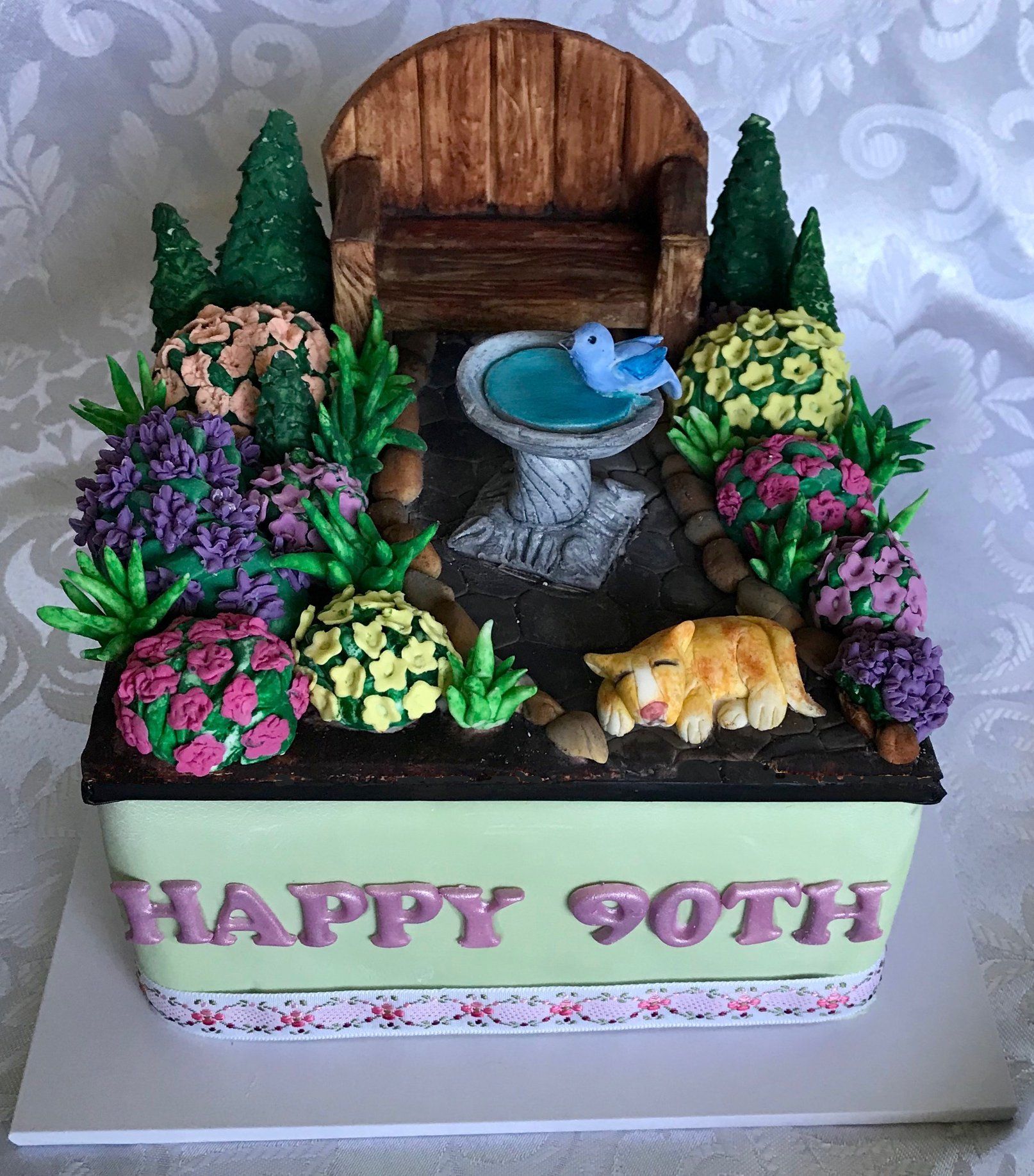 Square Garden themed birthday cake with garden chair, bird bath, flowering bushes and little cat.