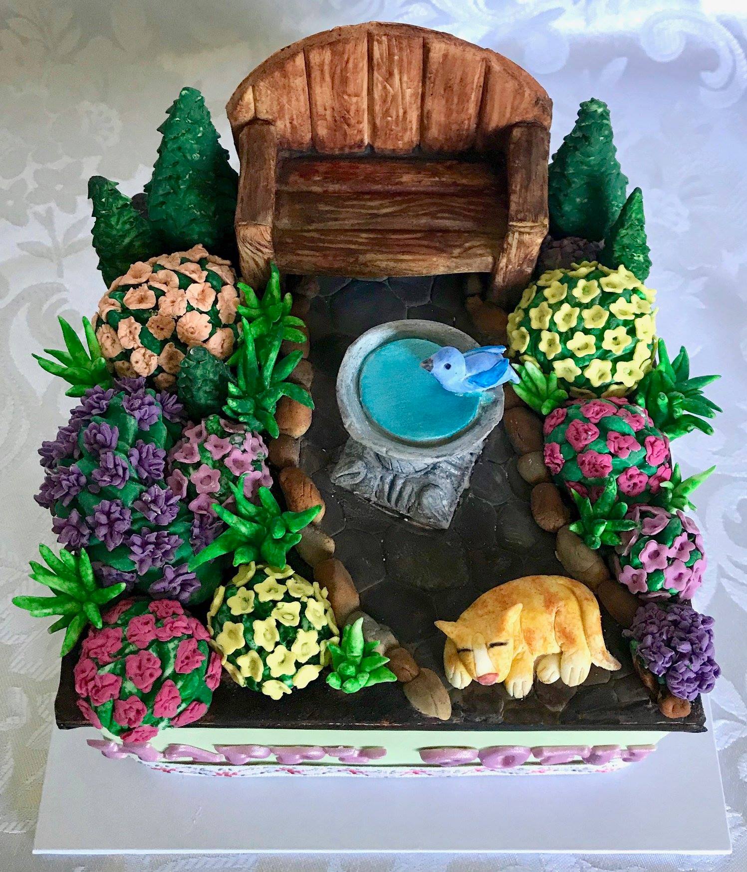 Square Garden themed birthday cake with garden chair, bird bath, flowering bushes and little cat.