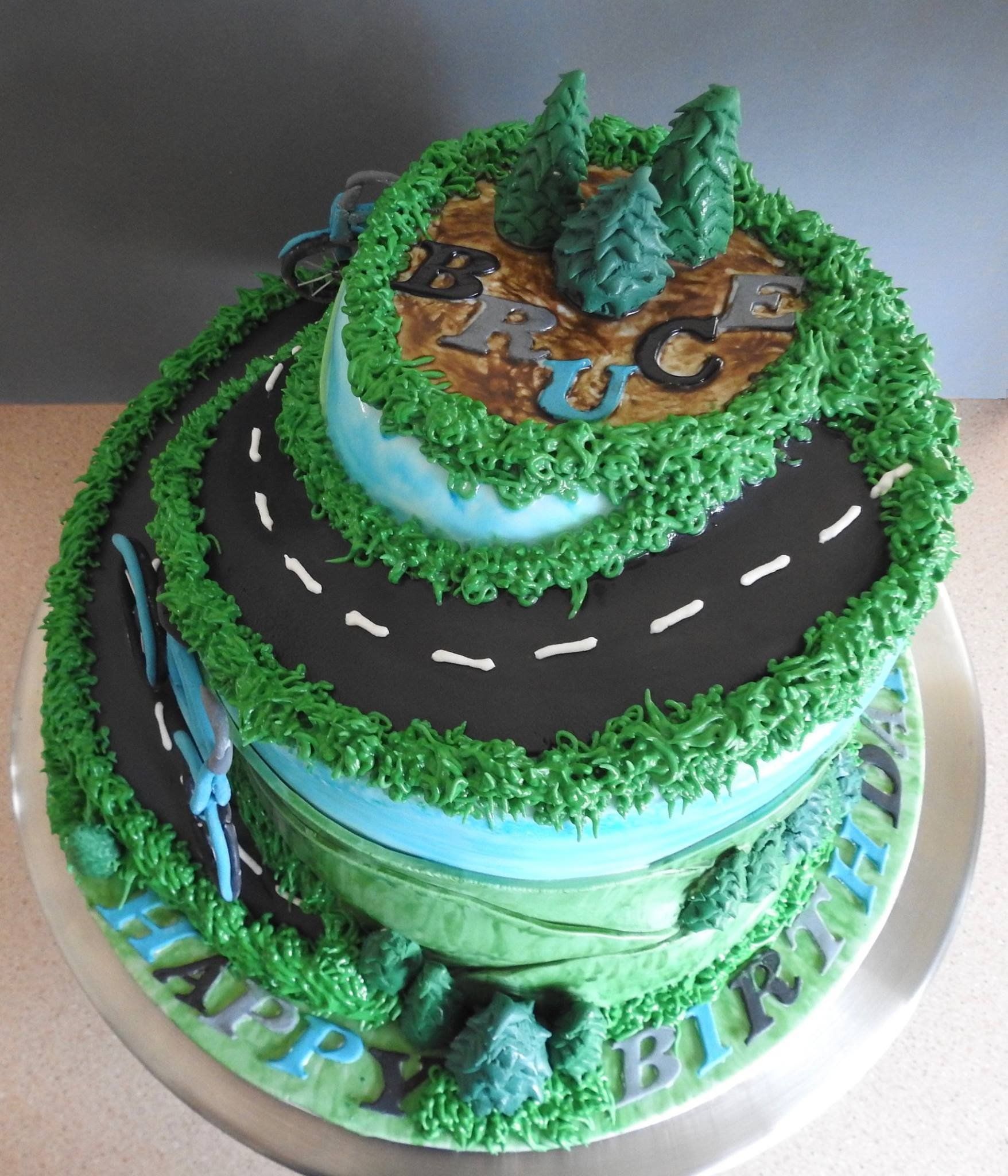 Tierd and shaped birthday cake with fondant hills, trees and two bicycles coming down a road.