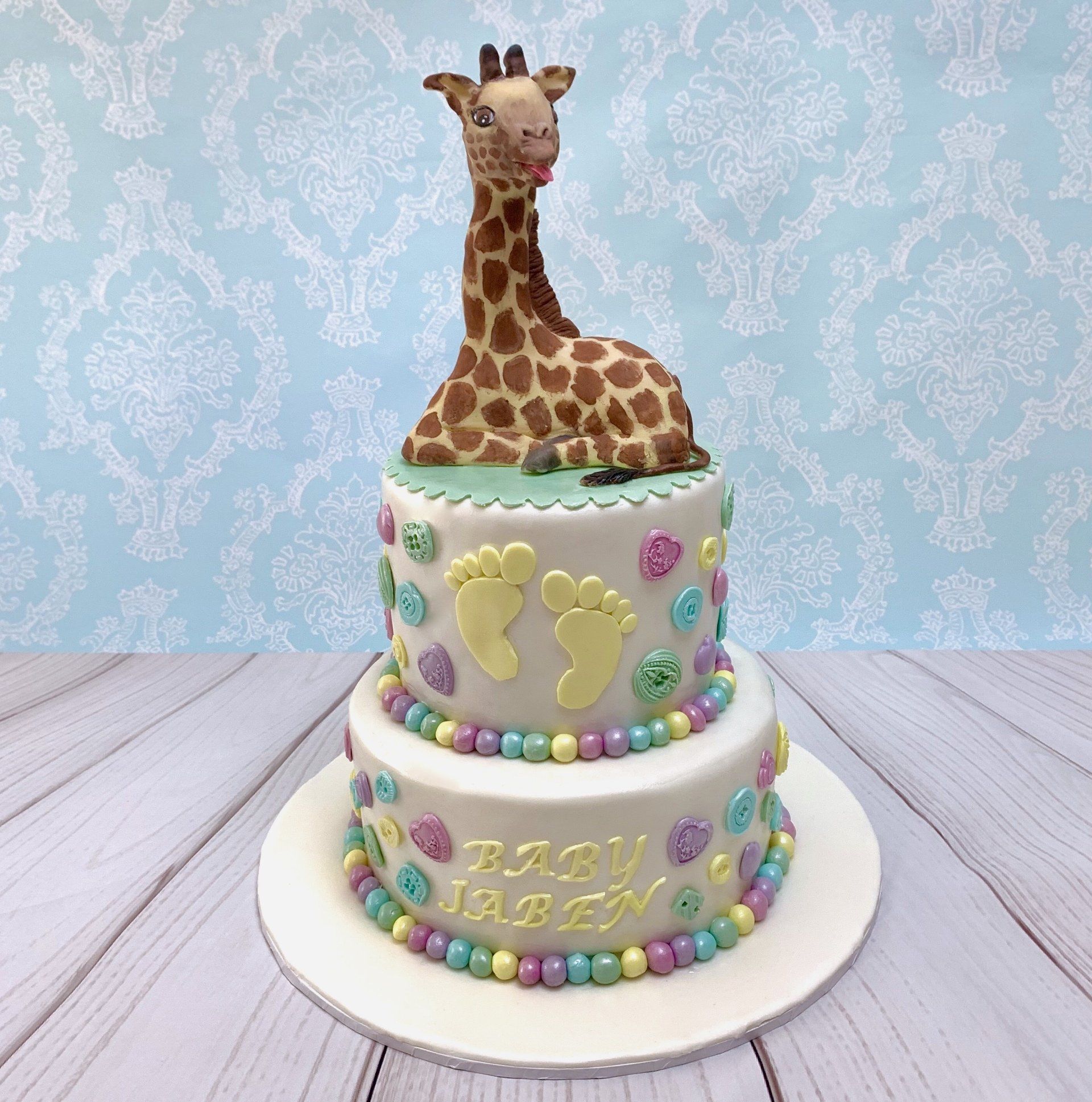 Two tie round baby shower cake. Cute Handsculpted fondant giraffe on top, little baby feet on the front of the top tier and pastel coloured fondant hearts and buttons on the sides of both tiers.