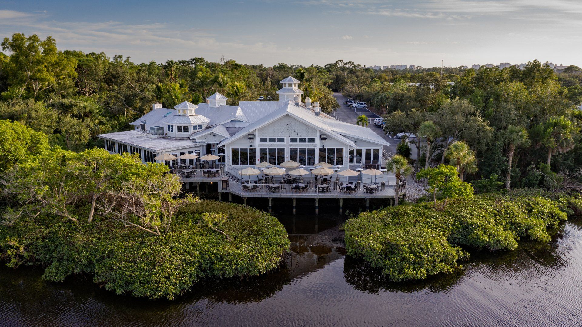 Large white waterfront house surrounded by lush green trees and calm dark water at sunset