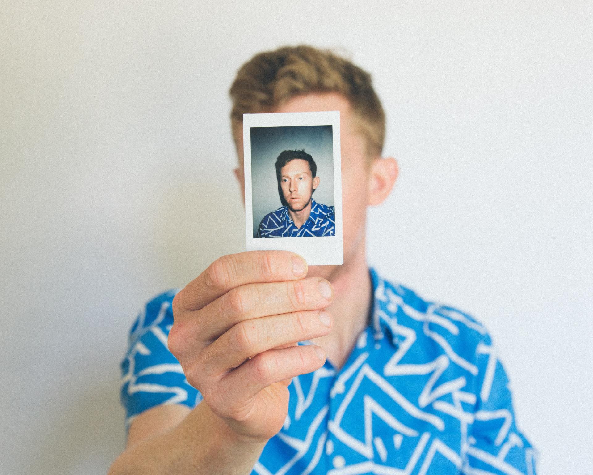 A man in a blue shirt is holding a polaroid picture in front of his face