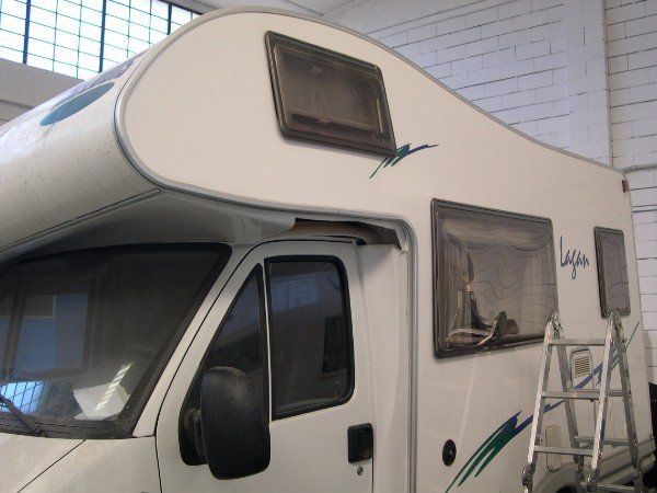 Un camper bianco con una piccola finestra e una scaletta aperta appoggiata ad essa, accanto a un muro di mattoni.
