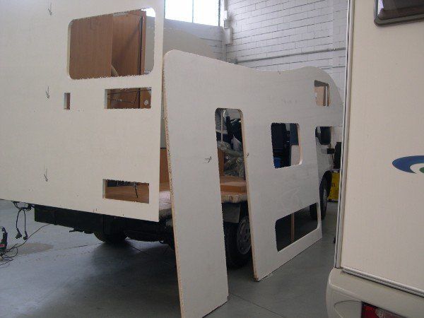 Camper o roulotte bianca con il pannello laterale rimosso, che lascia intravedere l'interno, e un pannello staccato appoggiato accanto ad esso.