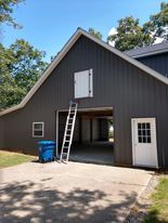 Gray barn with open garage, white door, ladder, and blue trash can on concrete.