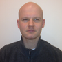 A bald man wearing a black jacket looks at the camera