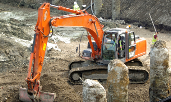 construction site mechanical excavator working around exposed concrete foundation piles preparing basement earthworks